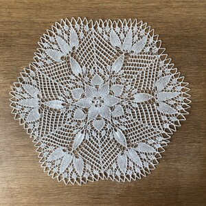 Könnte beinhalten: Weiße gehäkelte Tischdecke mit einem zarten Blumenmuster. Die Tischdecke ist rund und hat einen gewellten Rand.