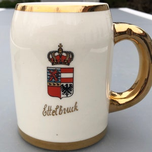Op de afbeelding: Witte keramische bierkan met een gouden rand en handvat. De kan heeft een wapenschild met een kroon, een schild met een rood-wit ontwerp en de tekst "Ettelbruck".