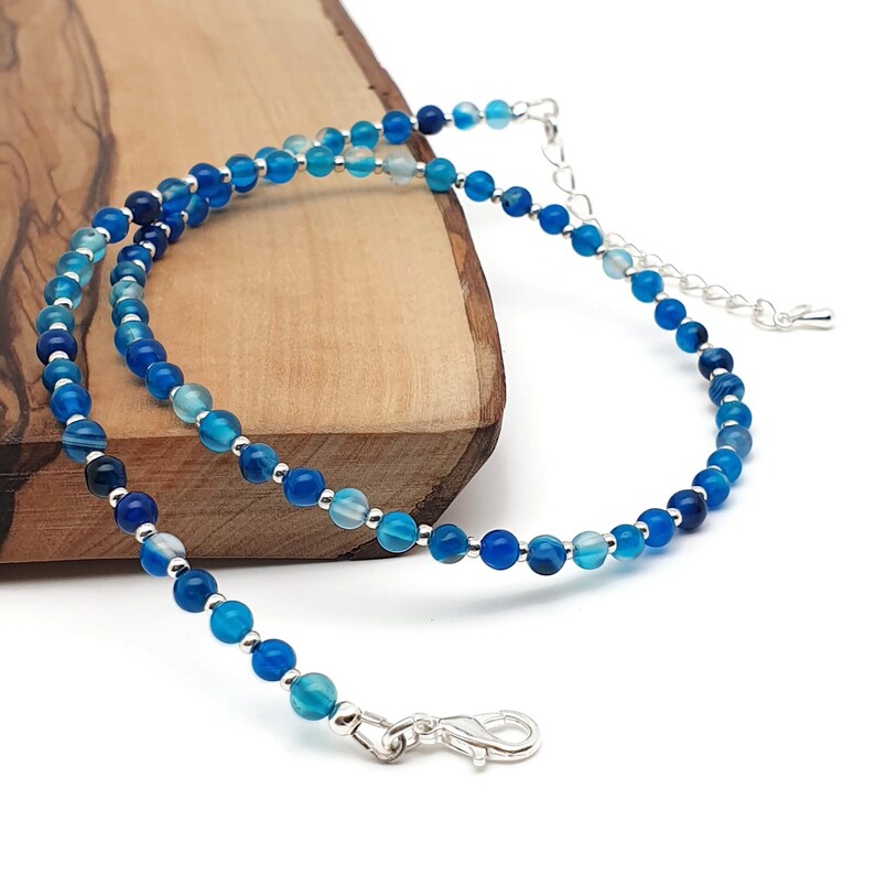 Beaded blue Agate necklace, made with 4 mm round gemstone beads interspaced with 2 mm silver plated glass beads, strung on flexible silicon plated steel beading wire. Finished with silver plated brass lobster claw clasp and extension chain. On the edge of a wooden board.