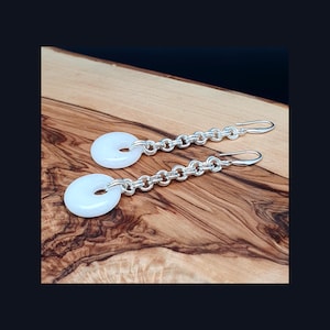 May include: A pair of earrings with a white, circular stone pendant with a hole in the middle, attached to a silver chain and hook. The earrings are displayed on a wooden surface.