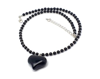 Op de afbeelding: Een zwarte kralenketting met een hartvormige hanger. De ketting heeft kleine, ronde zwarte kralen geregen met een zilverkleurige ketting en sluiting. De hartvormige hanger is glad, glanzend zwart.