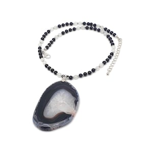 May include: A necklace featuring a large, oval-shaped agate pendant with a white and gray pattern. The pendant is suspended from a beaded chain with alternating black and white beads. The necklace has a silver-colored clasp and extender chain.
