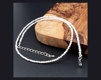 Op de afbeelding: Een delicate ketting bestaande uit kleine, ronde, wit en grijs gemarmerde kralen. Het sieraad is voorzien van een zilverkleurige karabijnsluiting en een verstelbare verlengketting met een kleine druppelvormige bedel aan het uiteinde.