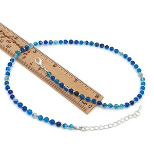 Beaded blue Agate necklace, made with 4 mm round gemstone beads interspaced with 2 mm silver plated glass beads, strung on flexible silicon plated steel beading wire. Finished with silver plated brass lobster claw clasp and extension chain. On top of a wooden ruler.