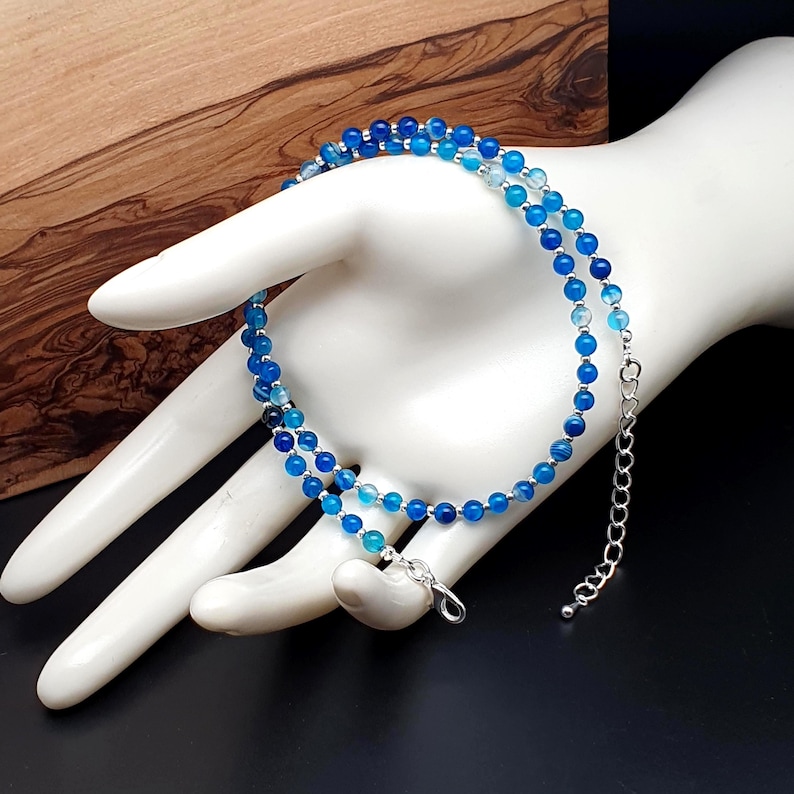 Beaded blue Agate necklace, made with 4 mm round gemstone beads interspaced with 2 mm silver plated glass beads, strung on flexible silicon plated steel beading wire. Finished with silver plated brass lobster claw clasp and extension chain. In white display hand prop with black and wooden background.