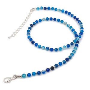 Beaded blue Agate necklace, made with 4 mm round gemstone beads interspaced with 2 mm silver plated glass beads, strung on flexible silicon plated steel beading wire. Finished with silver plated brass lobster claw clasp and extension chain. Coiled, with view of the clasp.