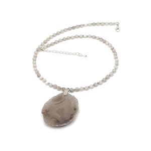 May include: A gray beaded necklace with a large, irregularly shaped, gray pendant. The necklace has a silver clasp and a delicate chain for adjusting the length. The pendant is a natural stone with a unique pattern.