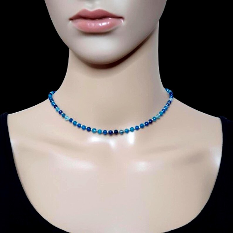 Beaded blue Agate necklace, made with 4 mm round gemstone beads interspaced with 2 mm silver plated glass beads. Finished with silver plated brass lobster claw clasp and extension chain. On a display bust with black background.