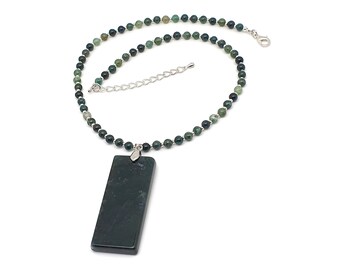 Op de afbeelding: Een ketting met een lange, rechthoekige, donkergroene stenen hanger aan een streng van kleine, ronde, groen en zwart gemêleerde kralen. De ketting is voorzien van een zilverkleurige karabijnsluiting en een verstelbare kettingverlenger.