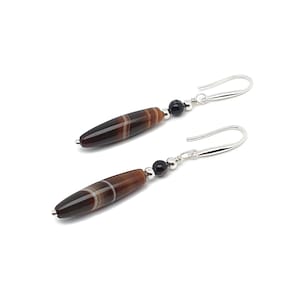 May include: A pair of dangle earrings with elongated, brown and white striped beads. Each earring has a small black bead and a silver-coloured hook. The earrings are set against a white background.