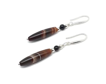 Op de afbeelding: Een paar oorhangers met langwerpige, bruin en wit gestreepte kralen. Elke oorbel heeft een kleine zwarte kraal en een zilverkleurige haak. De oorbellen staan tegen een witte achtergrond.
