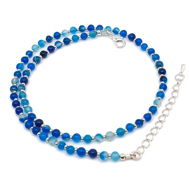 Beaded blue Agate necklace, made with 4 mm round gemstone beads interspaced with 2 mm silver plated glass beads, strung on flexible silicon plated steel beading wire. Finished with silver plated brass lobster claw clasp and extension chain. Coiled, with view of the chain.
