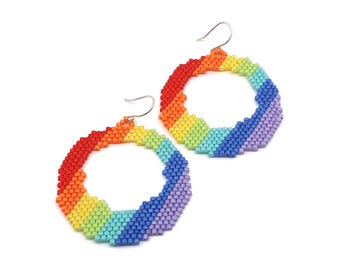 Op de afbeelding: Een paar ronde oorringen gemaakt van kleine, veelkleurige kralen die in een diagonaal regenboogpatroon zijn gerangschikt. De oorbellen bevatten een spectrum van rode, oranje, gele, groene, lichtblauwe, donkerblauwe en paarse kralen met zilverkleurige haken.