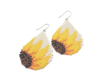 Puede incluir: Un par de pendientes en forma de lágrima hechos con pequeñas cuentas. El diseño presenta un patrón de girasol con un centro marrón oscuro, pétalos de color amarillo brillante y un fondo blanco. Cada pendiente cuelga de un gancho de metal plateado.