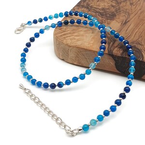 Beaded blue Agate necklace, made with small round gemstone beads interspaced with tiny silver plated glass seed beads, strung on flexible silicon plated steel beading wire. Finished with silver plated brass lobster claw clasp and extension chain. On the edge of a wooden board.