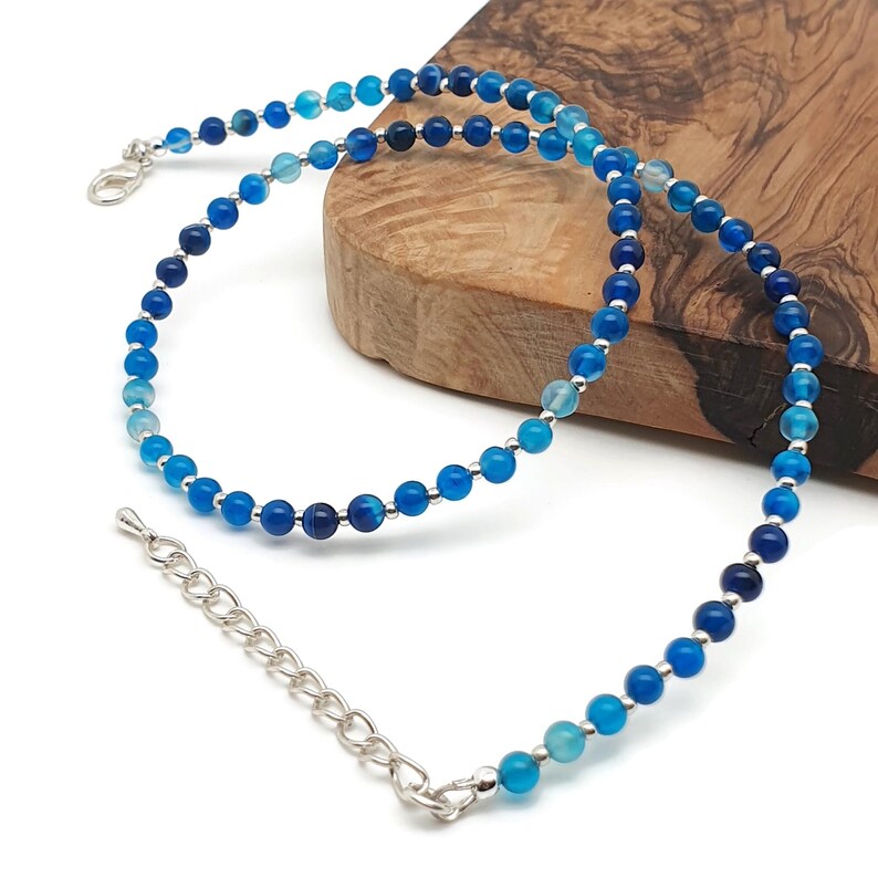Beaded blue Agate necklace, made with small round gemstone beads interspaced with tiny silver plated glass seed beads, strung on flexible silicon plated steel beading wire. Finished with silver plated brass lobster claw clasp and extension chain. On the edge of a wooden board.