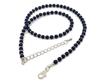 Op de afbeelding: Een ketting bestaande uit een doorlopende reeks kleine, ronde, gepolijste zwarte kralen afgewisseld met kleine zilverkleurige metalen tussenstukjes. Het sieraad is voorzien van een zilverkleurige karabijnsluiting en een verlengketting.