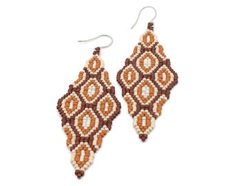 Op de afbeelding: Een paar ruitvormige bungelende oorbellen gemaakt van kleine kraaltjes in tinten bruin, oranje en crème. De oorbellen hebben een herhalend geometrisch patroon en zijn bevestigd aan zilverkleurige metalen oorhaken om te dragen.