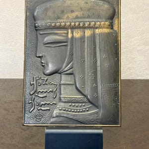 May include: A rectangular metal wall plaque featuring a dark, embossed profile of a person wearing a decorative headpiece and veil. The surface displays intricate dotted patterns and Georgian script text on the left side. The plaque is mounted on a dark base.