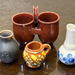 以下が含まれることがあります： 葉の彫刻が施されたダークグレーのジャグ、2つの取っ手が付いた茶色のマグカップ、黄色と白の花柄が描かれた小さなオレンジ色のポット、青い花が描かれた白いピッチャーを含む、4つのミニチュア陶器容器のコレクション。