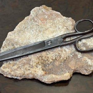 Pode incluir: Uma tesoura de metal cinza-escuro com dois anéis ovais e um parafuso central. A tesoura apresenta lâminas longas e retas com um acabamento texturizado e desgastado, repousando sobre uma formação rochosa cristalina de cor castanho-claro.