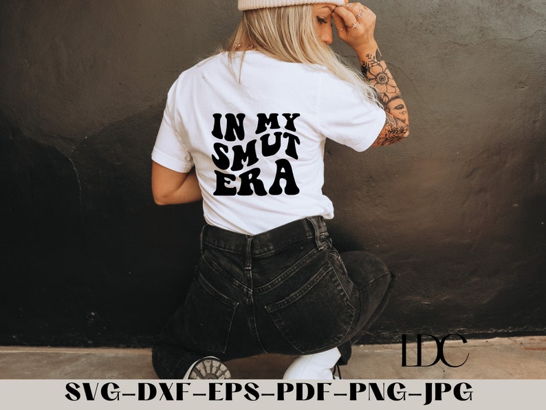 May include: A white t-shirt with black text that reads "IN MY SMUT ERA". The shirt is being worn by a person with tattoos on their arm.