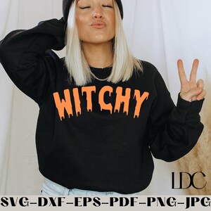 以下が含まれることがあります： 黒いスウェットシャツに、オレンジ色の垂れ下がった文字で「Witchy」とプリントされています。スウェットシャツは、長いブロンドの髪と黒いビーニー帽をかぶった人が着用しています。