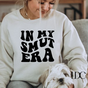 May include: A white sweatshirt with black text that reads "In My Smut Era" in a retro font. The sweatshirt is being worn by a person and a small white dog is sitting on their lap.