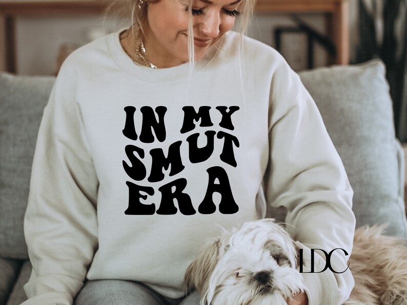 May include: A white sweatshirt with black text that reads "In My Smut Era" in a retro font. The sweatshirt is being worn by a person and a small white dog is sitting on their lap.