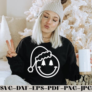 May include: A black sweatshirt with a white design of a smiling face wearing a Santa hat with two Christmas trees for eyes. The person wearing the sweatshirt is holding a white mug.
