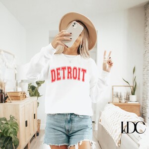 May include: White sweatshirt with red lettering that says "DETROIT" in a collegiate font. The sweatshirt is being worn by a person with a brown hat and a peace sign.