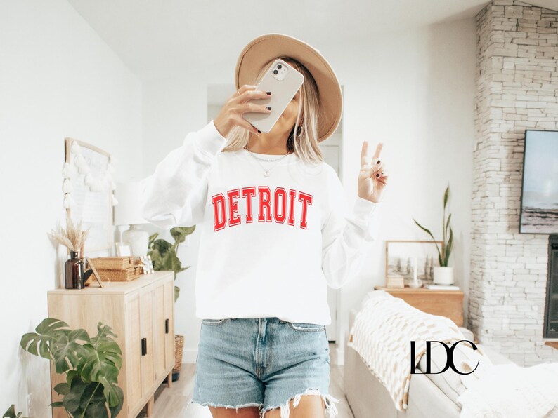 May include: White sweatshirt with red lettering that says "DETROIT" in a collegiate font. The sweatshirt is being worn by a person with a brown hat and a peace sign.