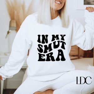 May include: White sweatshirt with black text that reads "In My Smut Era" in a retro font.