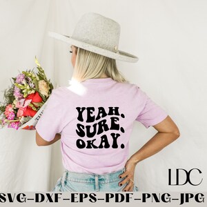 Pode incluir: Uma camiseta rosa com o texto "YEAH. SURE. OKAY." em letras pretas de estilo retrô. A camiseta está sendo usada por uma pessoa com um chapéu cinza claro.