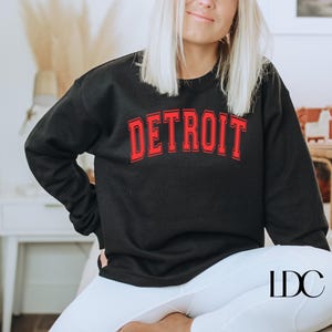 May include: A black sweatshirt with a red "DETROIT" graphic on the front. The sweatshirt is being worn by a person with long blonde hair. The person is sitting down with their legs crossed.