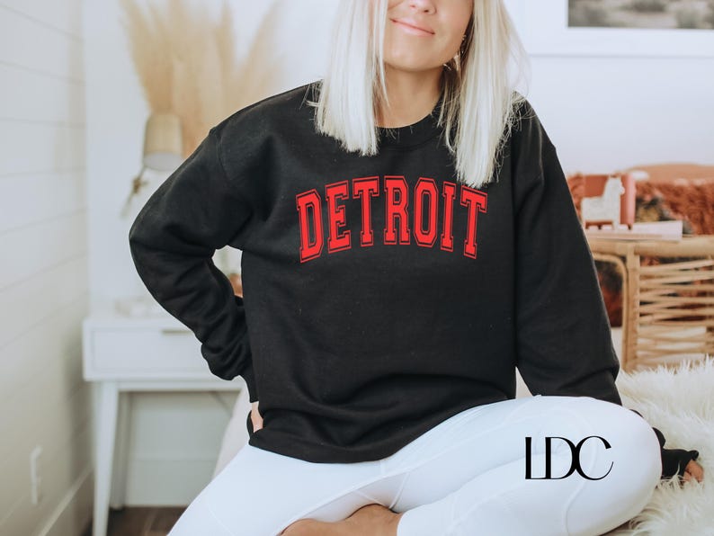 May include: A black sweatshirt with a red "DETROIT" graphic on the front. The sweatshirt is being worn by a person with long blonde hair. The person is sitting down with their legs crossed.