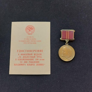 May include: A gold-colored circular medal featuring a profile portrait of Vladimir Lenin with the dates 1870-1970, attached to a red ribbon bar with yellow stripes. Beside it is a beige document with red Cyrillic text and a Soviet emblem.