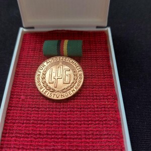 Puede incluir: Una medalla circular de color bronce con las letras LPG en el centro, rodeada por una corona y el texto FÜR AUSGEZEICHNETE LEISTUNGEN. La medalla está sujeta a una pequeña cinta verde, negra y amarilla y descansa dentro de un estuche blanco.