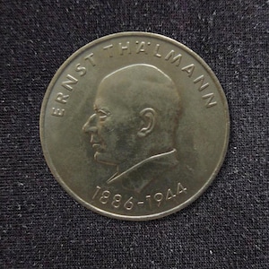 Könnte beinhalten: Eine kreisförmige Metallmünze mit einem Profilporträt von Ernst Thälmann, das nach links gerichtet ist. Der Text ERNST THÄLMANN ist am oberen Rand eingeprägt, und die Jahreszahlen 1886-1944 sind am unteren Rand der Münzoberfläche eingeprägt.