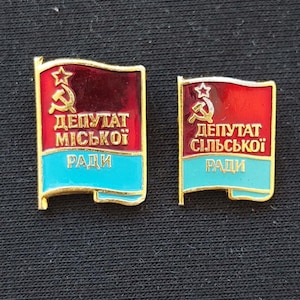 May include: Two rectangular metal pins shaped like waving flags with gold trim. The left pin features a red field with a gold hammer and sickle and star, and text reading DEPUTAT MISKOI RADY above a light blue stripe. The right pin is similar with text reading DEPUTAT SILSKOI RADY.