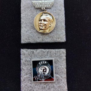 Op de afbeelding: Twee metalen pins op grijze viltvierkanten. De bovenste pin is een cirkelvormig goudkleurig medaillon met het portret van een man, hangend aan een trapeziumvormige balk met de tekst ZVEZDNY GORODOK. De onderste pin is een vierkant met een zwart-rode afbeelding van een astronaut in een helm met de tekst CCCP.