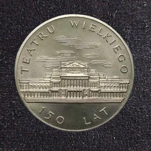 May include: A round silver-toned commemorative coin featuring an embossed architectural building in the center. The text TEATRU WIELKIEGO is arched along the top, and the text 150 LAT is positioned along the bottom edge of the coin face.