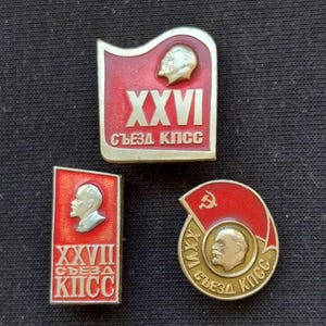 May include: Three Soviet-era metal lapel pins featuring red enamel and gold-tone accents. One rectangular pin displays XXVI СЪЕЗД КПСС, a second rectangular pin shows XXVII СЪЕЗД КПСС, and a circular pin features a hammer and sickle with XXVI СЪЕЗД КПСС.
