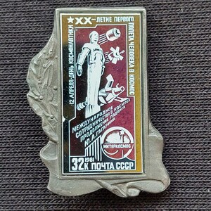 May include: A rectangular metal pin featuring a space-themed graphic with a white figure of a cosmonaut, a satellite, and Cyrillic text. The design includes a red and black background, a star, and the text 32K POCHTA SSSR 1981, with a silver border.