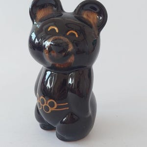 May include: A glossy dark brown ceramic bear figurine with rounded ears and a smiling face. The bear features light brown painted details on the ears and muzzle, and a gold-colored graphic of five interlocking rings around its midsection.