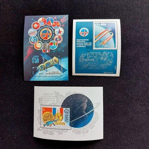 May include: Three rectangular postage stamps featuring space exploration themes. One stamp shows a space station and circular mission emblems. Another displays a rocket and the text POLSKA. The third features a stylized astronaut silhouette.