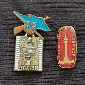 May include: Two metal pins from East Germany. One pin features a blue flag with the letters FDJ above a rectangular plaque depicting the Berlin TV Tower and the word BERLIN. The second pin is oval, red, and gold, showing the TV Tower with the text BERLIN HAUPTSTADT DER DDR.