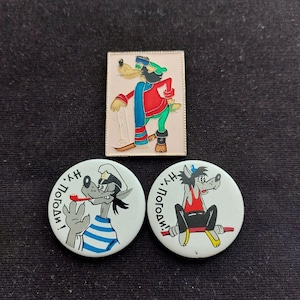May include: A rectangular metal pin featuring a cartoon wolf on skis, alongside two circular pinback buttons. One button shows a wolf in a sailor shirt with a pipe, and the other shows a wolf sitting on a red bench, both with the text "Ну, погоди!".