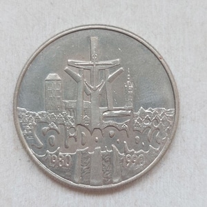 May include: A silver-coloured commemorative coin featuring a central cross monument, a cityscape silhouette, and the text Solidarność. The dates 1980 and 1990 are embossed at the bottom of the circular coin, which has a smooth, metallic finish.