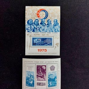 Op de afbeelding: Een herdenkingsvel met postzegels van de Apollo-Sojoez-missie uit 1975 met portretten van vijf astronauten, het missielogo en een blauwe postzegel met een controlecentrum. Daaronder drie postzegels met scènes van maanonderzoek.